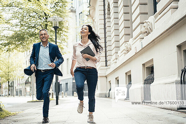 Cheerful businessman and businesswoman running on footpath in city
