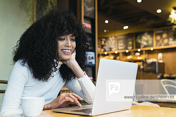 Smiling young woman with hand on chin using laptop at cafe