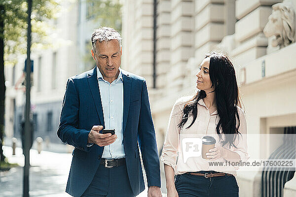 Businesswoman holding disposable coffee cup walking with businessman using smart phone on footpath