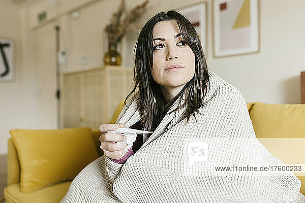 Beautiful woman wrapped in blanket holding thermometer in living room