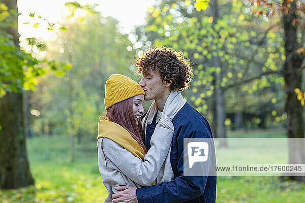 Young man embracing and kissing girlfriend's forehead in autumn park