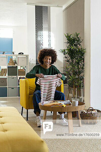 Smiling young Afro woman folding striped t-shirt sitting on chair at home