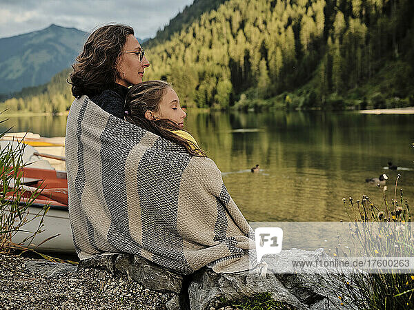 Mother and daughter wrapped in blanket sitting at lakeshore