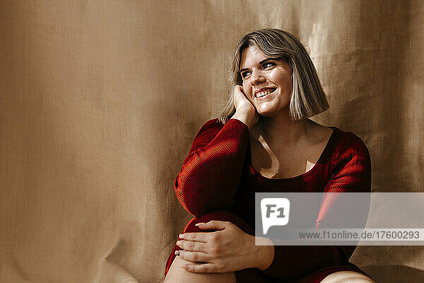 Smiling curvy woman with hand on cheek sitting in front of brown backdrop