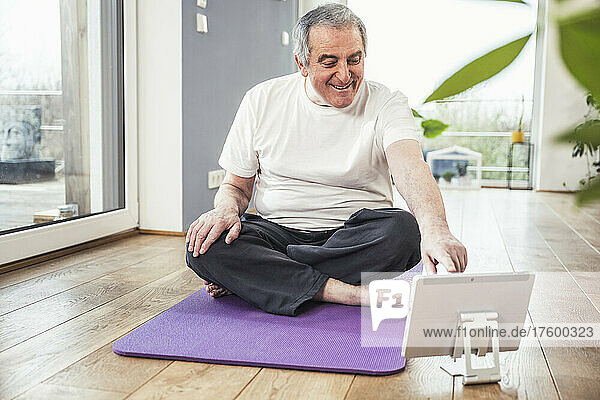Smiling senior man sitting cross-legged on exercise mat using tablet PC at home