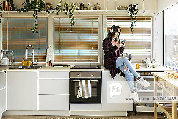 Woman with headphones using smart phone on kitchen counter at home