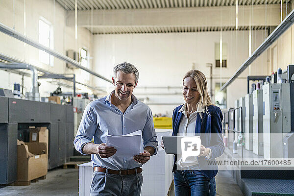 Smiling blond businesswoman with tablet PC standing by colleague reading document in factory