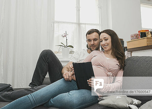 Lächelnde schwangere Frau teilt Tablet-PC mit Mann  der zu Hause auf dem Sofa liegt