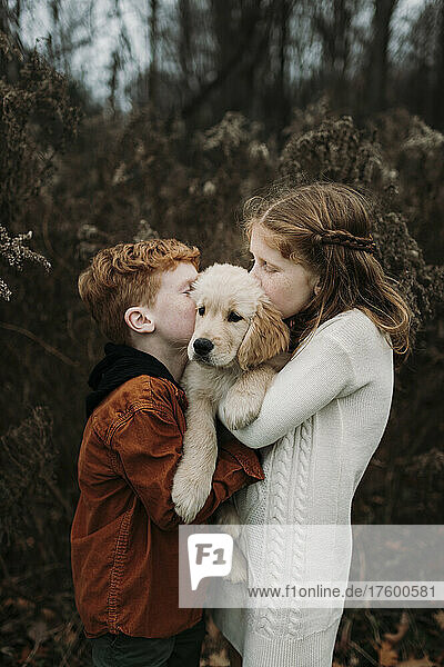 Brother and sister kissing golden retriever puppy in forest