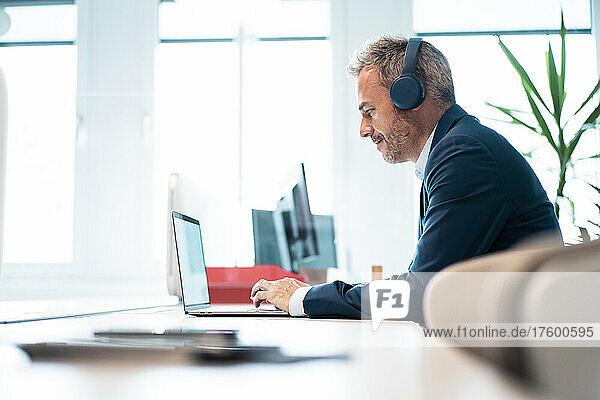 Businessman wearing wireless headphones using laptop at desk in workplace
