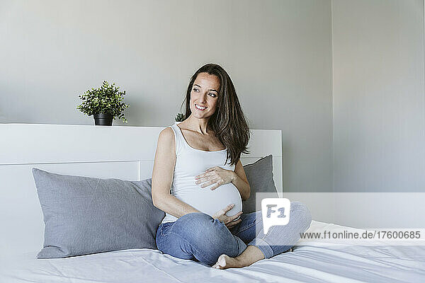 Happy pregnant woman with long brown hair sitting on bed at home