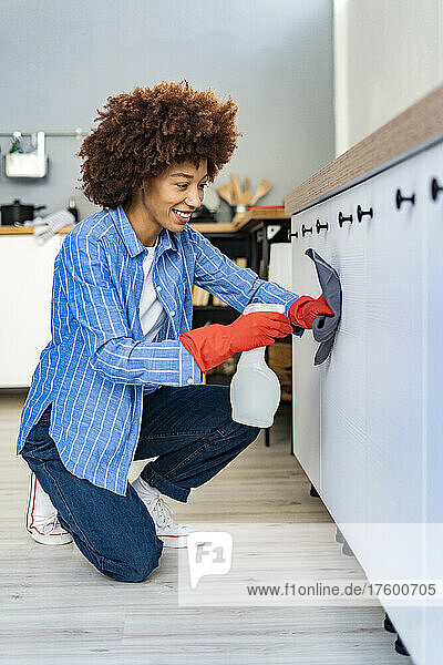 Smiling Afro woman kneeling and spraying disinfection on cabinet in kitchen