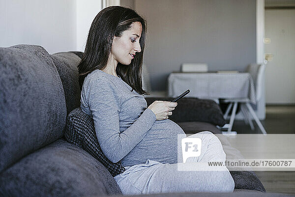 Pregnant woman using smart phone sitting on sofa at home