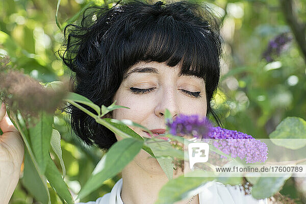 Smiling woman with closed eyes smelling purple flowers on plant