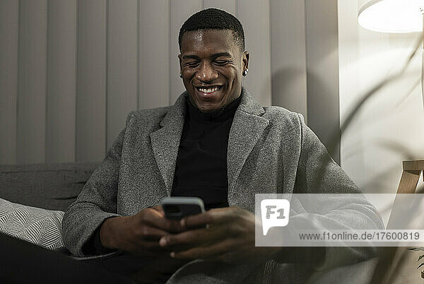 Smiling young man using smart phone sitting on sofa at home