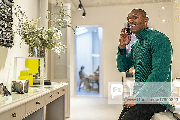 Glücklicher Mann im grünen Pullover am Telefon  der zu Hause am Schreibtisch steht