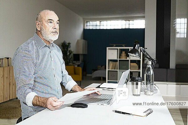 Thoughtful businessman with laptop and document sitting at desk in home office