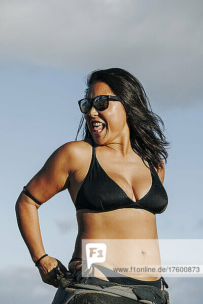 Cheerful woman in sunglasses wearing wetsuit and bikini at beach