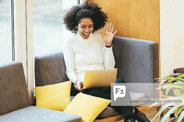 Cheerful businesswoman waving on video call through laptop on sofa
