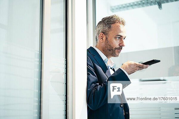 Businessman talking through smart phone speaker leaning on doorway at office