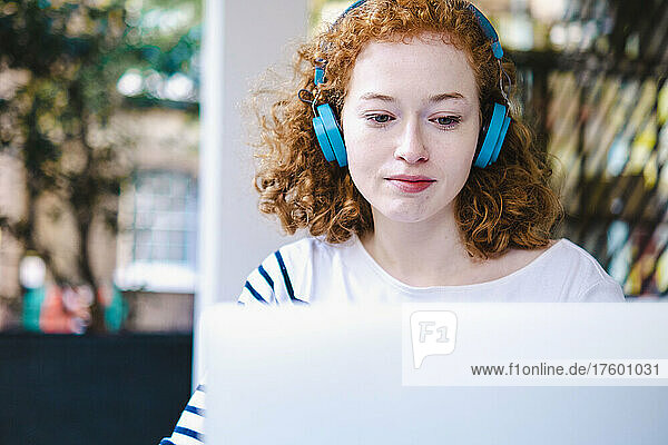 Redhead woman using laptop and listening music on headphones in cafe