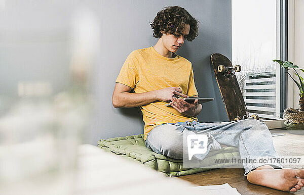 Junger Mann nutzt Tablet-PC mit Skateboard zu Hause