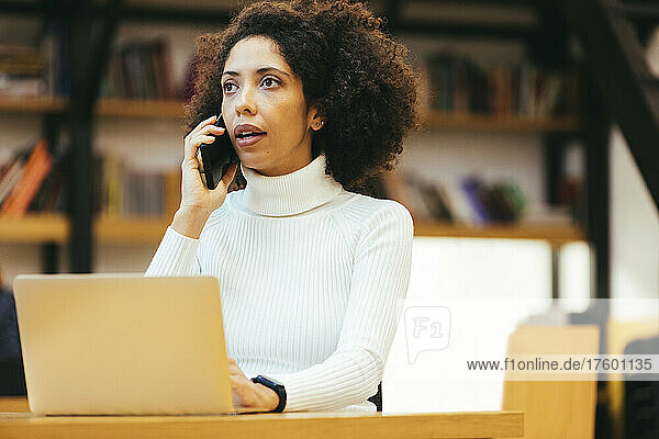 Businesswoman talking on smart phone with laptop at desk in office