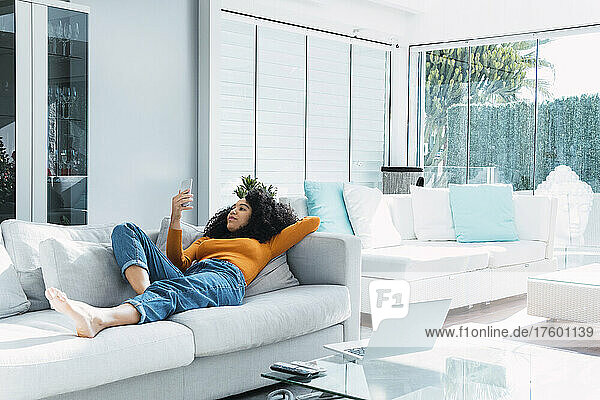 Woman using smart phone lying on sofa in living room