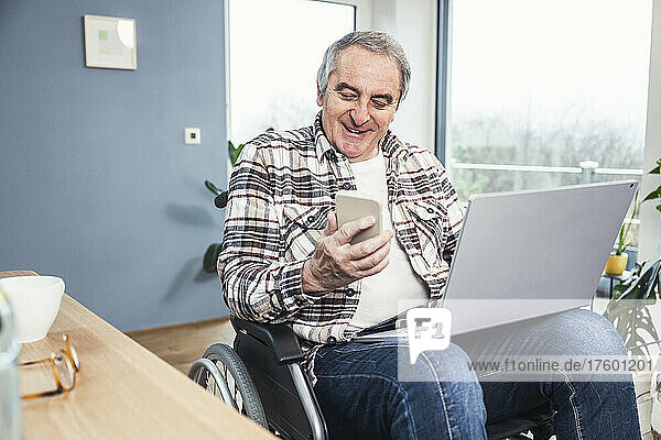 Smiling disabled man with laptop using smart phone on wheelchair at home