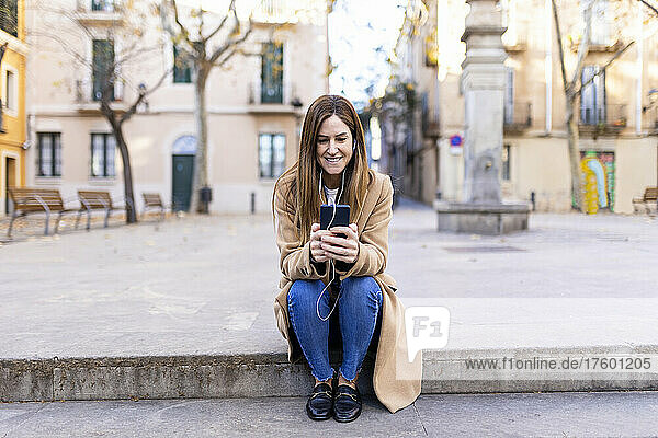 Lächelnde Frau  die auf dem Fußweg sitzt und mit ihrem Mobiltelefon im Internet surft