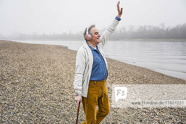 Happy senior man with hand raised listening music through headphones at beach