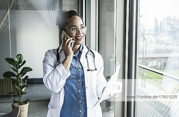 Lächelnder Arzt mit Dokument  der im Krankenhaus auf dem Smartphone spricht
