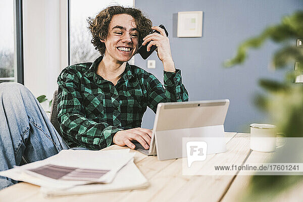 Happy young man talking on mobile phone through speaker sitting at table in living room