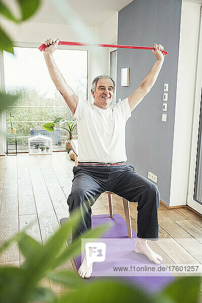 Smiling senior man with arms raised holding resistance band sitting on chair at home