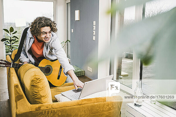 Smiling young man holding guitar using laptop on sofa at home