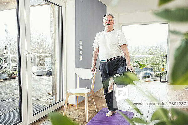 Smiling senior man standing on one leg by chair at home