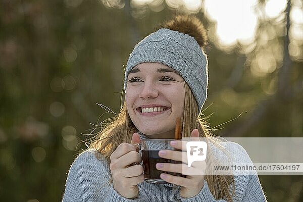 Portrait of a young woman in winter with cap  laughing  warming herself with a cup of tea  in the forest  Upper Bavaria  Bavaria  Germany  Europe