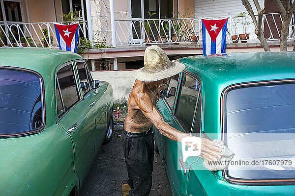 Mit kubanischen Flaggen in der Nähe wäscht ein älterer Mann mit einem Lappen einen amerikanischen Oldtimer in der Innenstadt von Havanna  Kuba; Havanna  Kuba