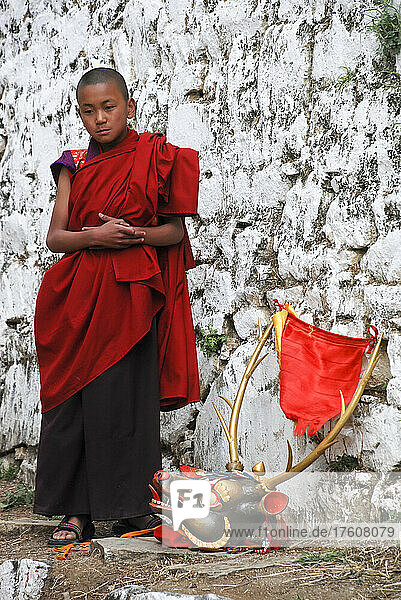 A young monk stands next to a mask used in the Paro Tshechu Festival within Paro Dzong  a monastery and fortress in Paro  Bhutan; Paro  Bhutan