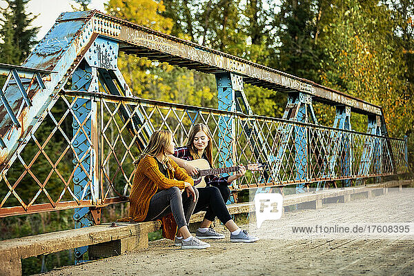 Two sisters sit on a park bridge  one with an acoustic guitar  enjoying music together; Edmonton  Alberta  Canada