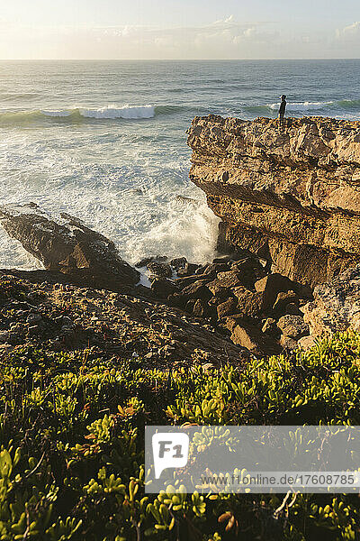 Eine Person steht auf einem zerklüfteten Bergrücken und blickt auf das Meer und die an die Küste rollenden Wellen; Praia do Guincho  Cascais  Portugal