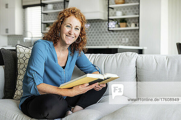A mid adult woman sits on a couch at home reading a book and looking at the camera; Edmonton  Alberta  Canada