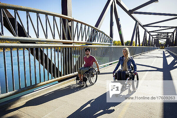 Young paraplegic man and woman going across a bridge together using their wheelchairs on a beautiful fall day; Edmonton  Alberta  Canada