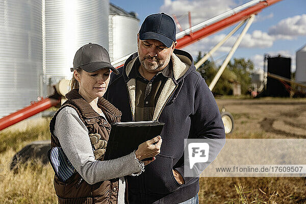 Mature couple working on their farm  standing in front of grain bins and auger and consulting their tablet computer; Alcomdale  Alberta  Canada