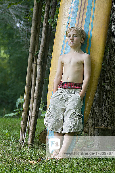 A six year old boy with boxers pulled high leans against a surfboard.; Cabin John  Maryland.