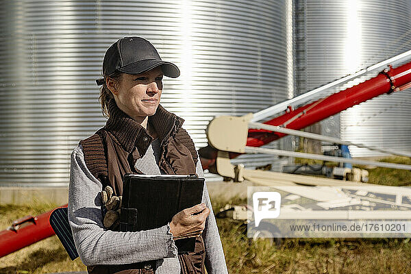 Porträt einer reifen Frau  die auf ihrer Farm arbeitet  einen Tablet-Computer in der Hand hält und vor Getreidebehältern und einer Schnecke steht; Alcomdale  Alberta  Kanada