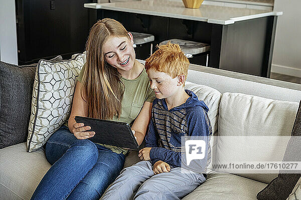 A brother and sister sit on a couch at home with a tablet and viewing something together; Alberta  Canada