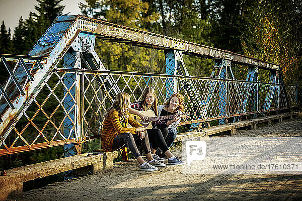 Mother and two sisters sit on a park bridge  one with an acoustic guitar  enjoying music together; Edmonton  Alberta  Canada