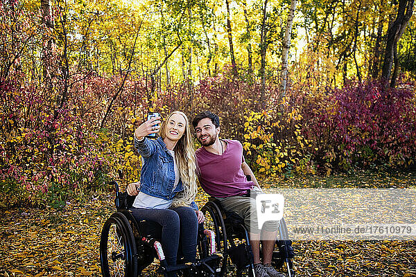 A young paraplegic man and woman in their wheelchairs taking a self portrait with a smart phone in a park on a beautiful fall day; Edmonton  Alberta  Canada