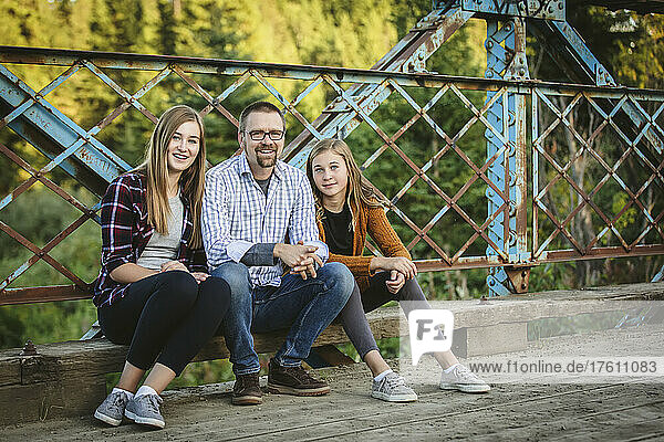 Outdoor portrait of a father with two teenage daughters sitting on a bridge in a city park in autumn; Edmonton  Alberta  Canada